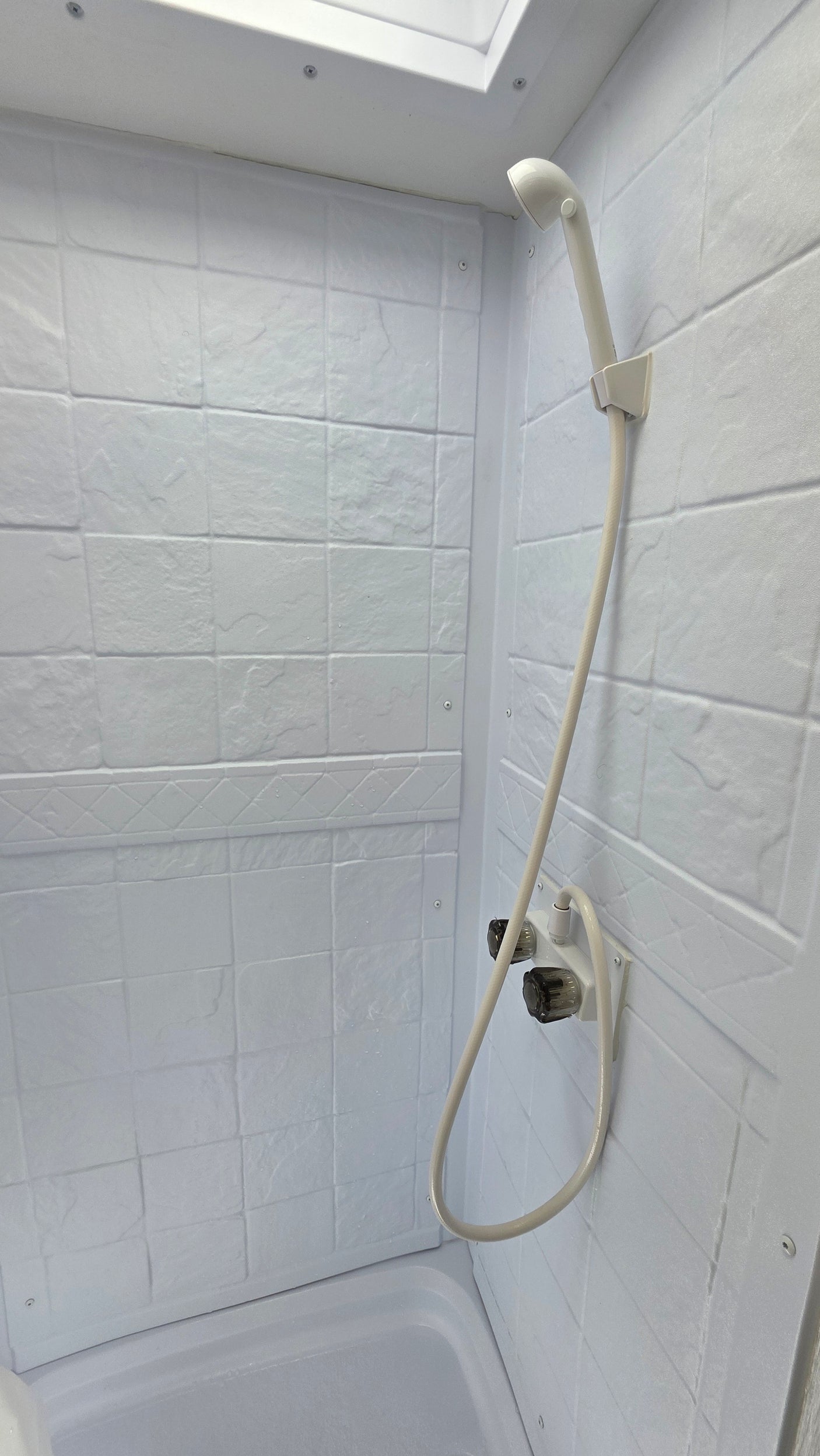 Clean, white shower stall in an RV rental, featuring a handheld showerhead, faucets, and textured tile-like walls.