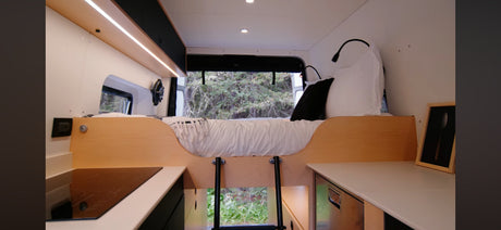RV rental interior: A compact camper van with a raised bed, ladder, modern kitchenette, and window views of nature.