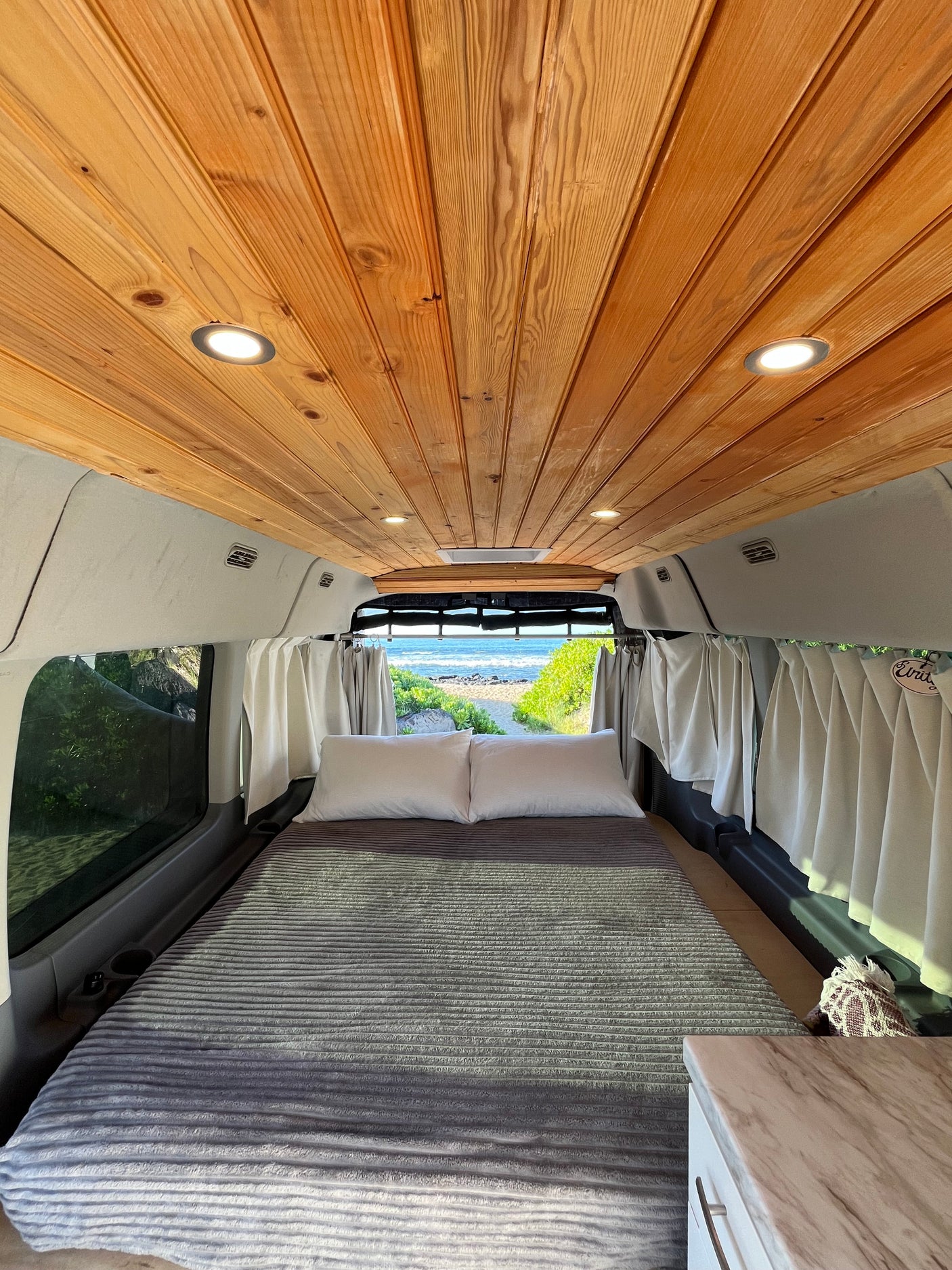 Inviting RV rental: cozy camper van interior with wood ceiling, comfy bed, and scenic beach view out the back.