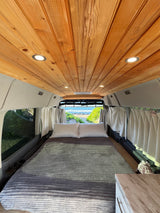 Inviting RV rental: cozy camper van interior with wood ceiling, comfy bed, and scenic beach view out the back.