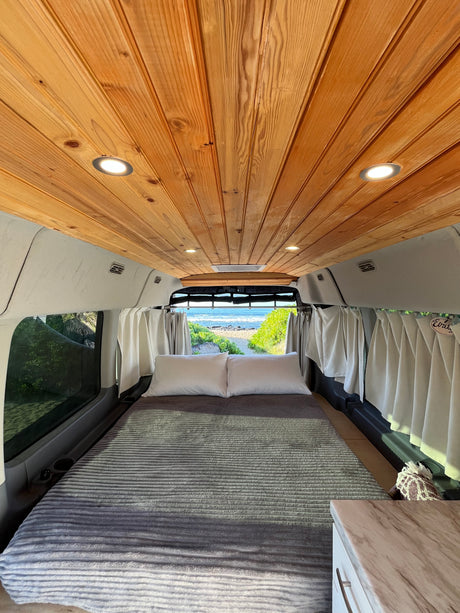 Inviting RV rental: cozy camper van interior with wood ceiling, comfy bed, and scenic beach view out the back.