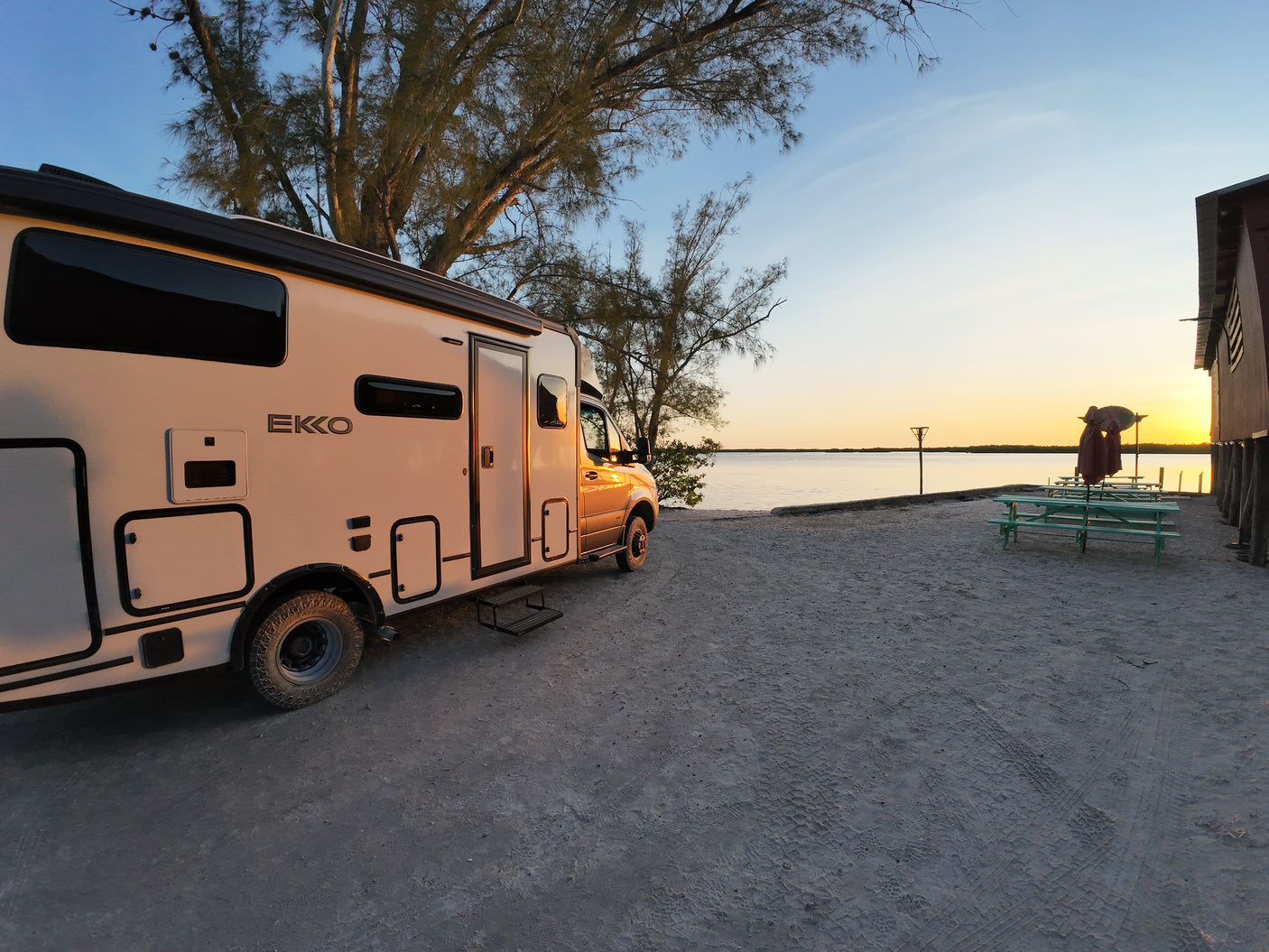 Waterfront RV rental offers stunning sunset views. This motorhome is parked on sandy ground by the bay at dusk.