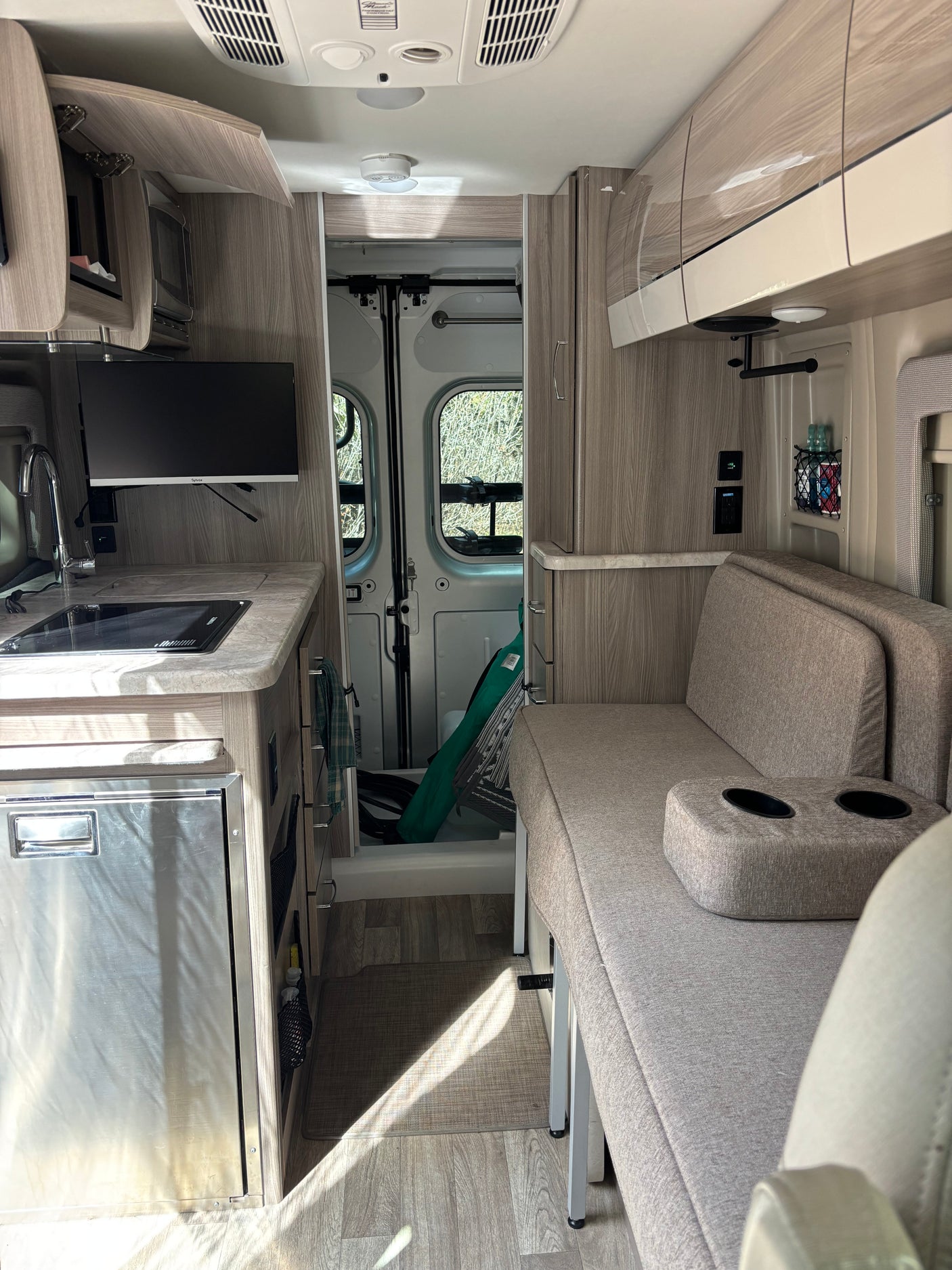 Inside a compact rv rental: kitchen with sink, stove, fridge, TV, & a sofa with cup holders. Back doors open to nature.
