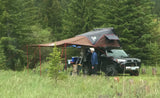 An SUV with a rooftop tent and awning, set up for overlanding, provides a unique RV rental experience. A man stands beside it.
