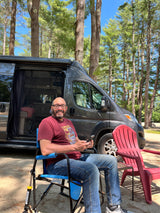 Man relaxing in a blue chair outside his gray RV rental at a sunny campground, surrounded by tall pine trees.