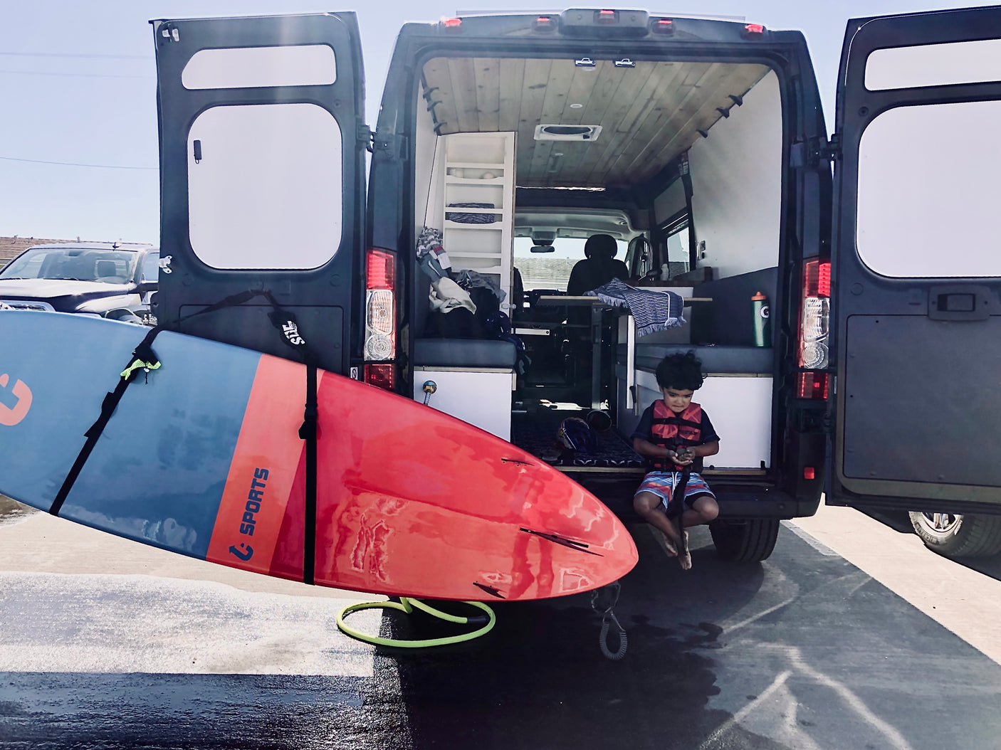Young child in a life vest sits at the back of an RV rental. A large paddleboard rests nearby, custom interior visible.