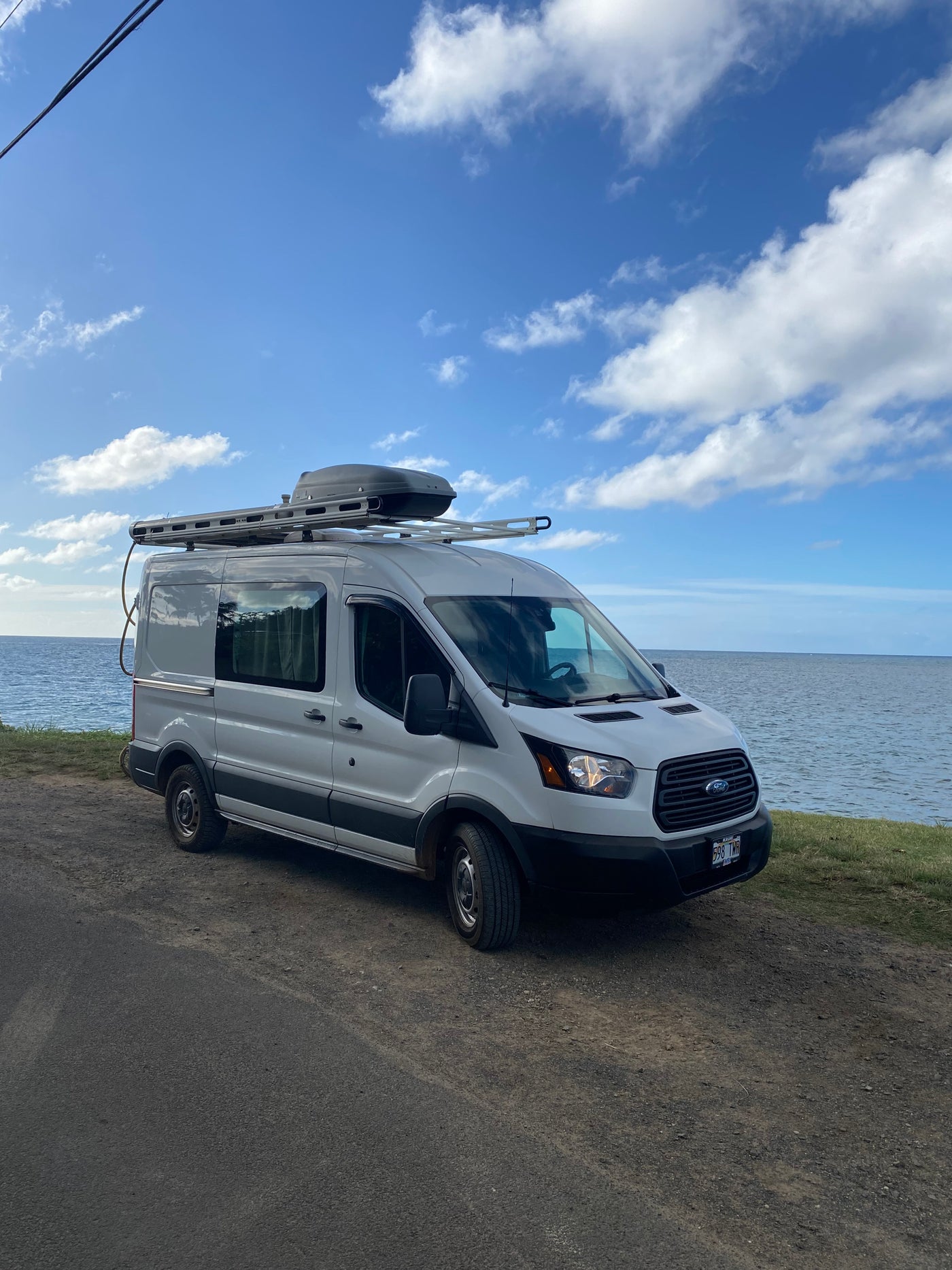 This white Ford Transit RV rental van is parked on a dirt road by the ocean under a beautiful blue, cloudy sky.
