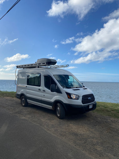 This white Ford Transit RV rental van is parked on a dirt road by the ocean under a beautiful blue, cloudy sky.