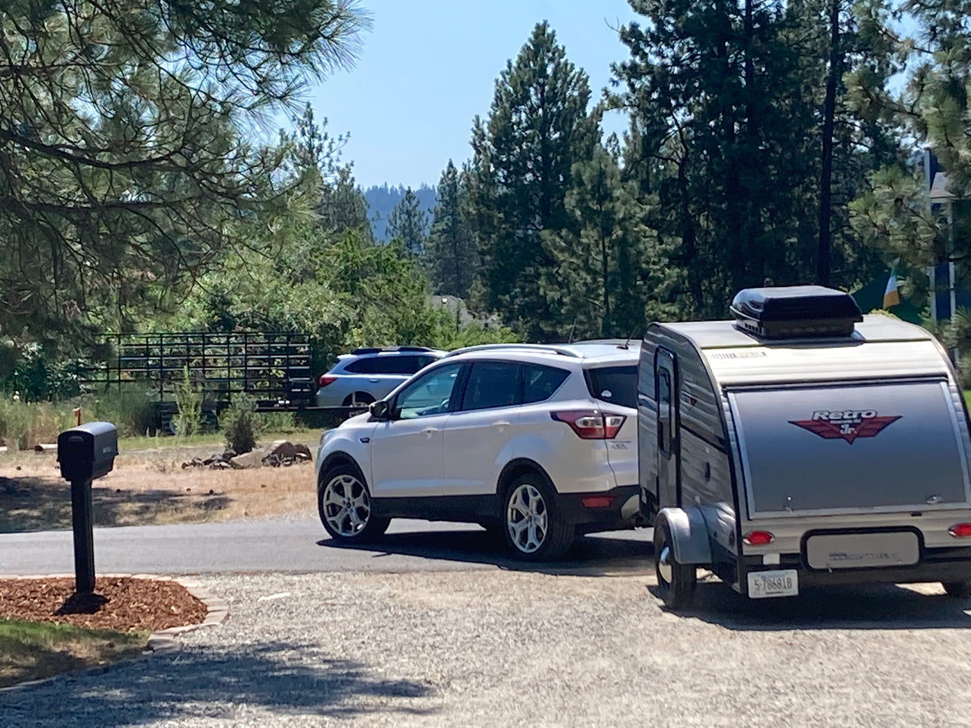 White SUV with a 'Retro Jr.' teardrop RV rental attached, parked on a sunny driveway near a mailbox and trees.