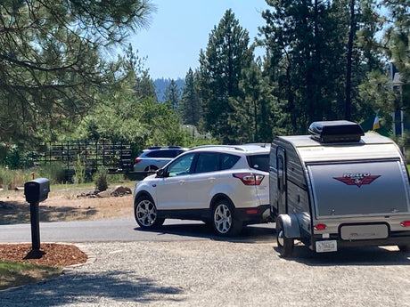 White SUV with a 'Retro Jr.' teardrop RV rental attached, parked on a sunny driveway near a mailbox and trees.