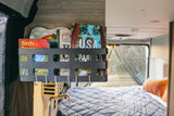 RV rental: Organized travel guides like "Moon USA" & "Birds of Texas" on a black wall rack, with a cozy bed inside.