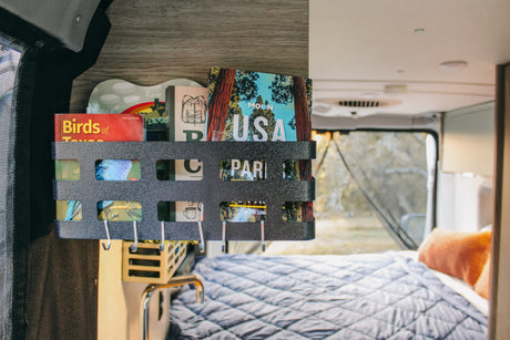 RV rental: Organized travel guides like "Moon USA" & "Birds of Texas" on a black wall rack, with a cozy bed inside.
