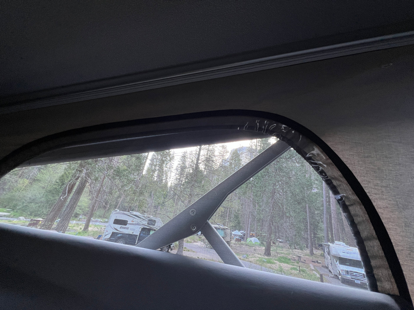 Looking out from an RV rental's screened window reveals a serene campground scene with tall trees, other RVs, and campers.
