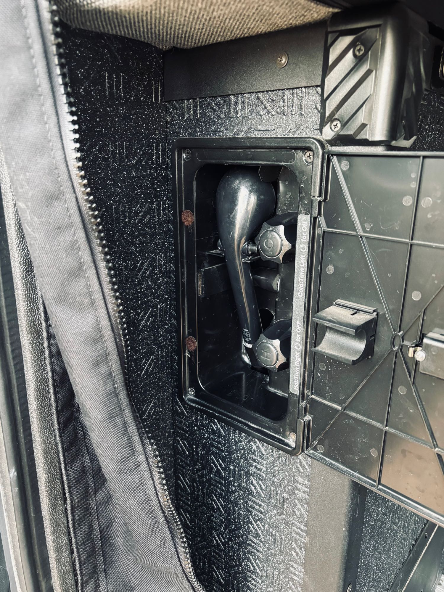 Compact outdoor shower system in an RV rental, showing a black shower head and hot/cold controls inside its compartment.