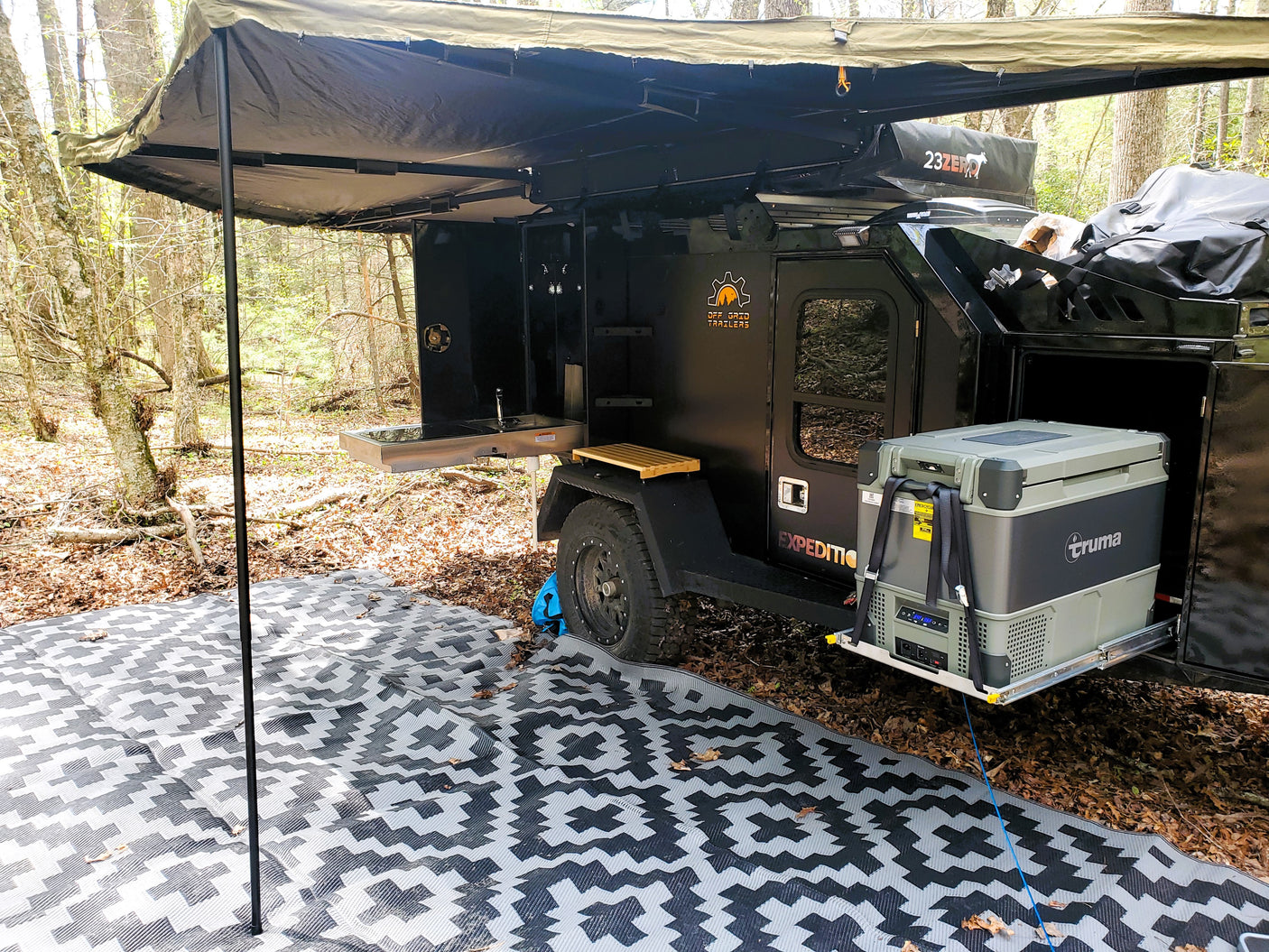 An RV rental overland trailer campsite with outdoor kitchen, Truma fridge, awning, and ground mat. Perfect for off-grid adventure!
