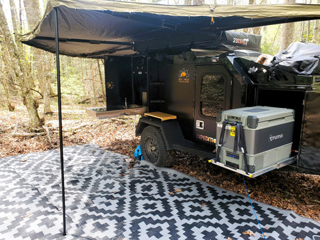 An RV rental overland trailer campsite with outdoor kitchen, Truma fridge, awning, and ground mat. Perfect for off-grid adventure!