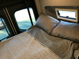 This RV rental shows a comfortable sleeping area with gray bedding and pillows, featuring large rear windows and a smaller camper window.