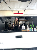 Woman in a versatile RV rental truck camper smiles, holding beer bottles. Interior shows a bed, drinks & tip jar.