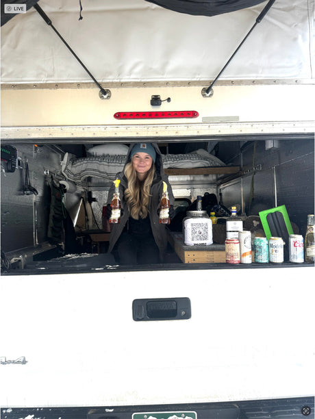 Woman in a versatile RV rental truck camper smiles, holding beer bottles. Interior shows a bed, drinks & tip jar.
