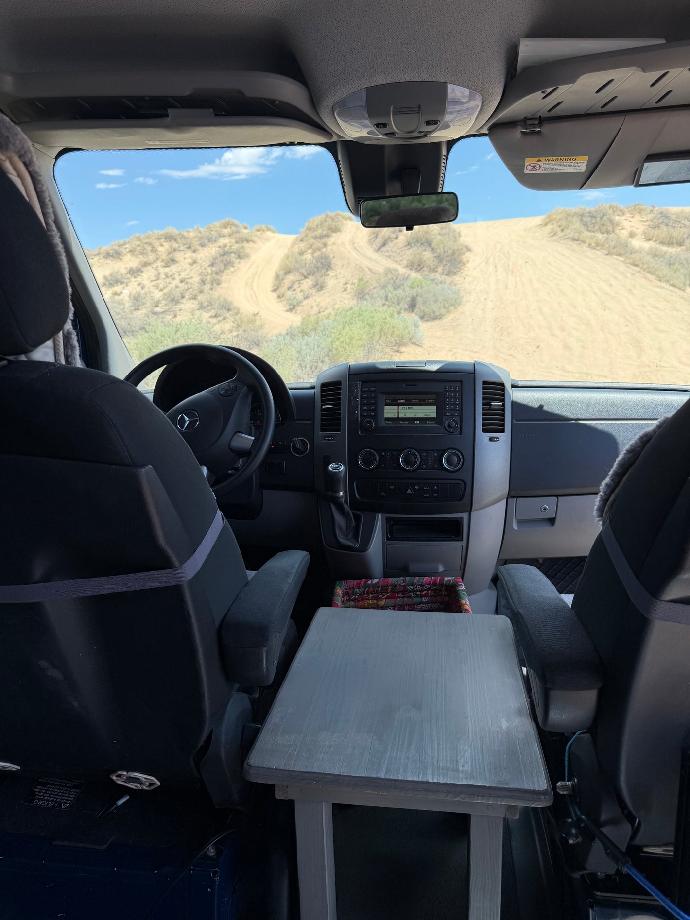 RV rental van cockpit view: Mercedes dashboard, steering wheel, and custom table, with a sandy desert trail outside. — photo 8