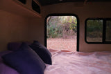 Inside an RV rental: a cozy bed with purple pillows and a view of autumn leaves through two windows.