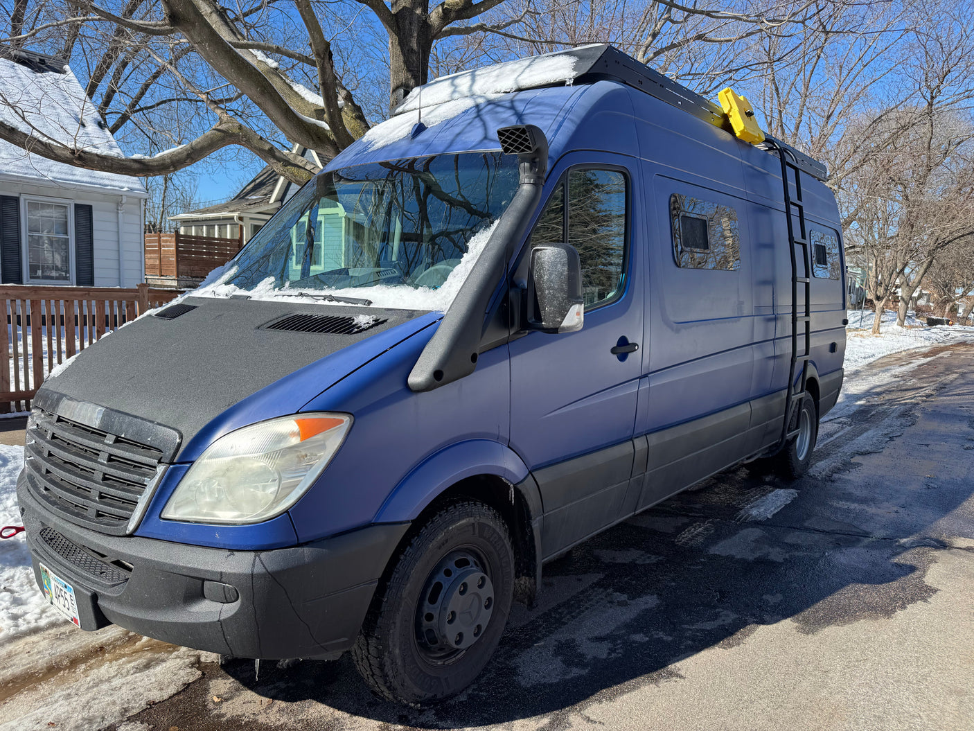 Custom blue Mercedes-Benz Sprinter camper van with snorkel, roof rack, and ladder, on a snowy street. Ideal RV rental.