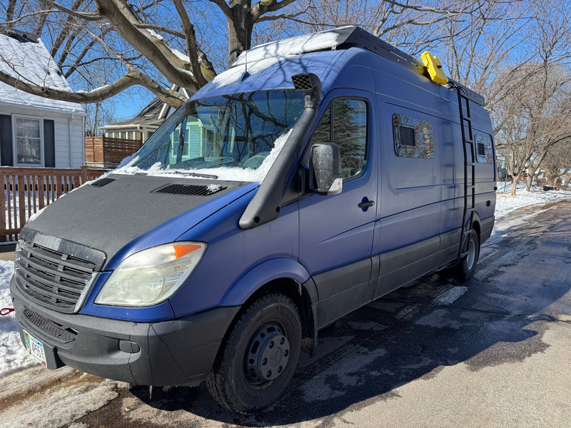 Custom blue Mercedes-Benz Sprinter camper van with snorkel, roof rack, and ladder, on a snowy street. Ideal RV rental. — photo 10