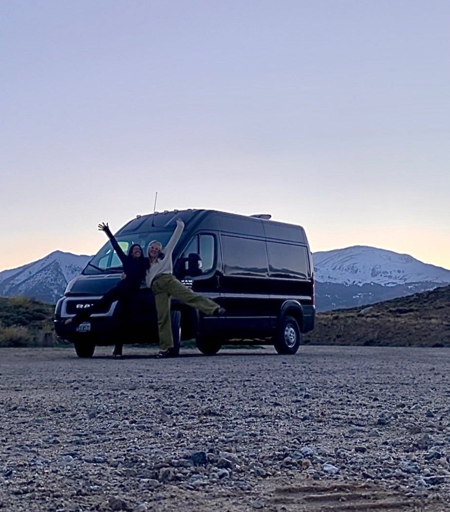 Two friends pose happily by their black Ram ProMaster RV rental van on a gravel lot, with snow-capped mountains in the background.