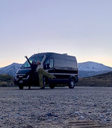 Two friends pose happily by their black Ram ProMaster RV rental van on a gravel lot, with snow-capped mountains in the background.