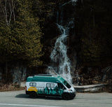 An RV rental camper van with mountain graphics is parked by a road, with a majestic waterfall cascading behind it.