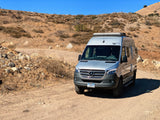 A silver Mercedes Sprinter camper van, an rv rental, parked on a dirt road amidst dry, rolling hills under a clear blue sky. Ready for travel.
