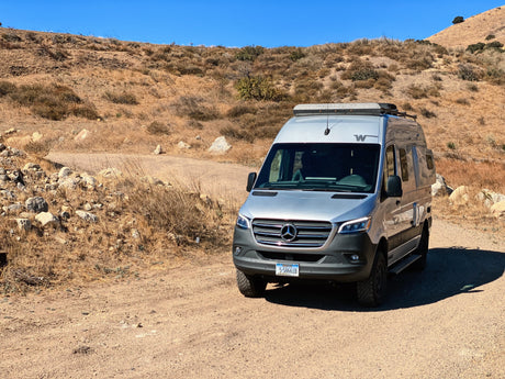 A silver Mercedes Sprinter camper van, an rv rental, parked on a dirt road amidst dry, rolling hills under a clear blue sky. Ready for travel.