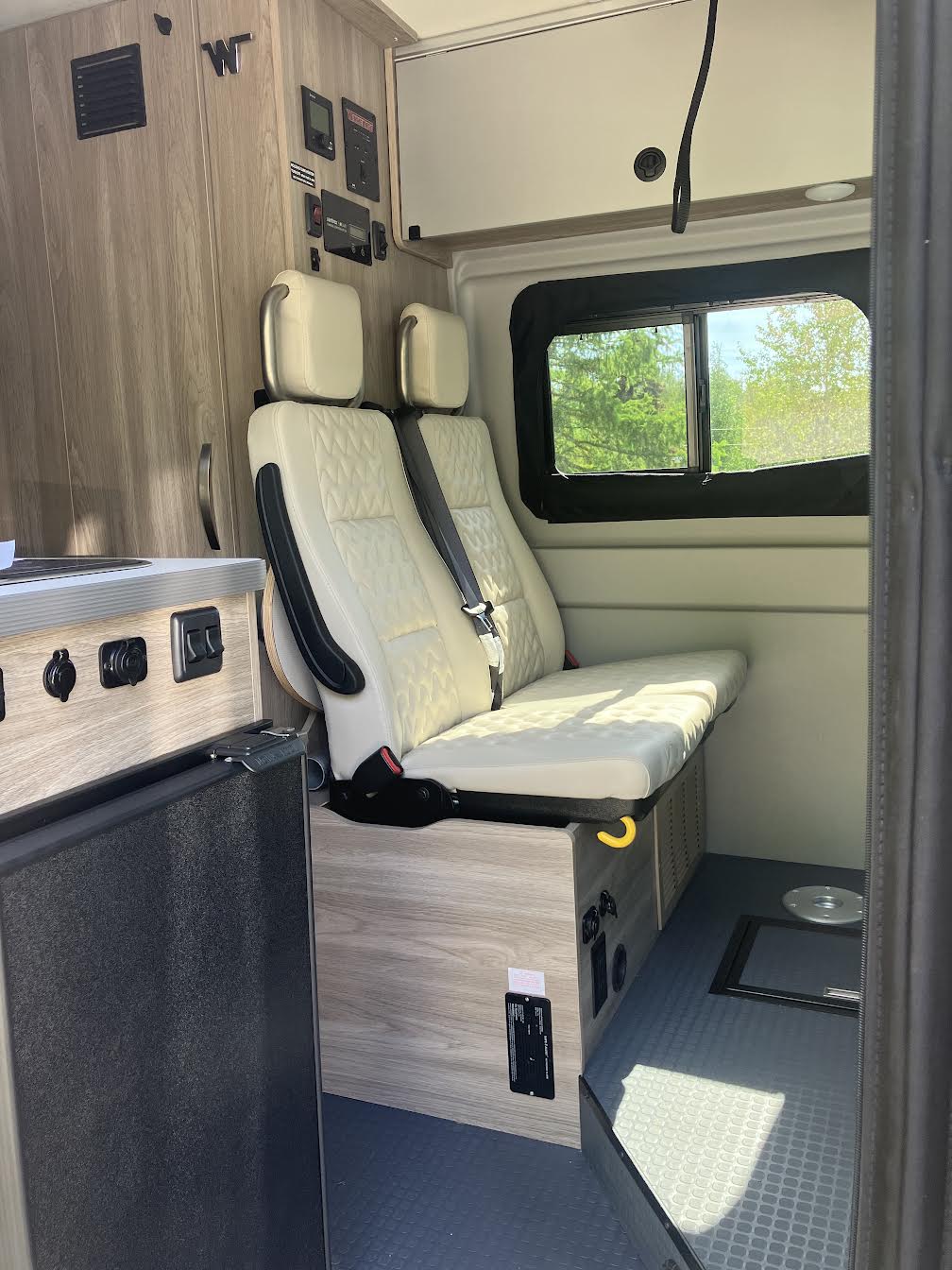 RV rental interior showcases two cream quilted seats converting to a bed, a fridge, control panels, and a window.