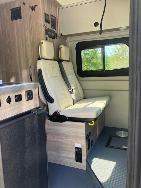 RV rental interior showcases two cream quilted seats converting to a bed, a fridge, control panels, and a window.