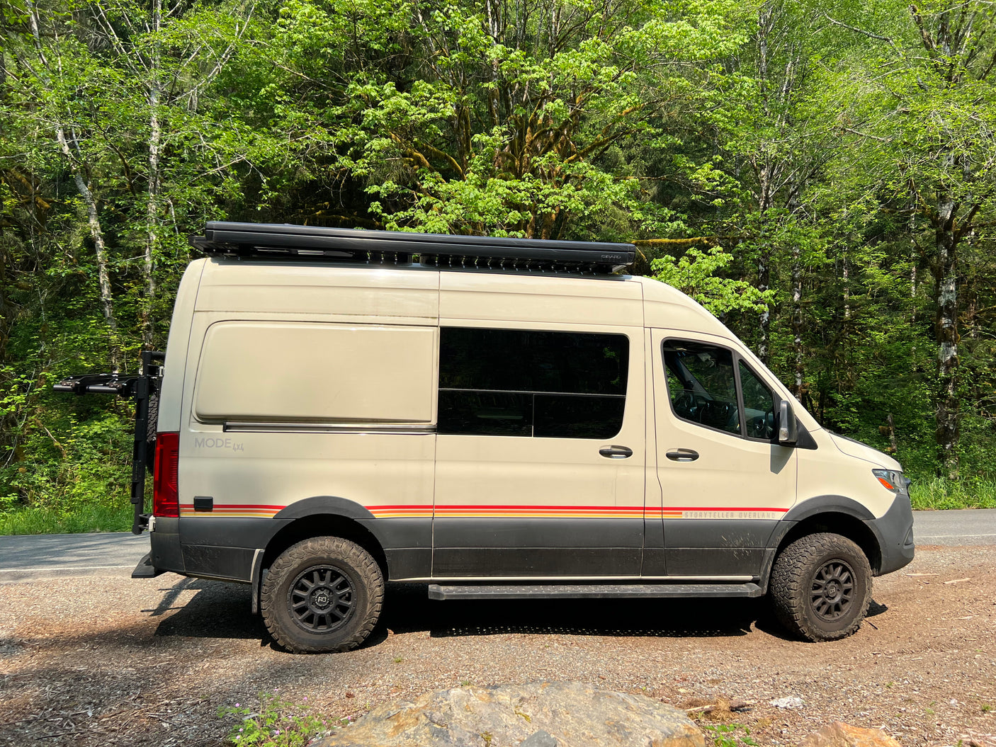 A cream-colored Storyteller Overland 4x4 camper van, ideal for RV rental adventures, parked by a lush forest.