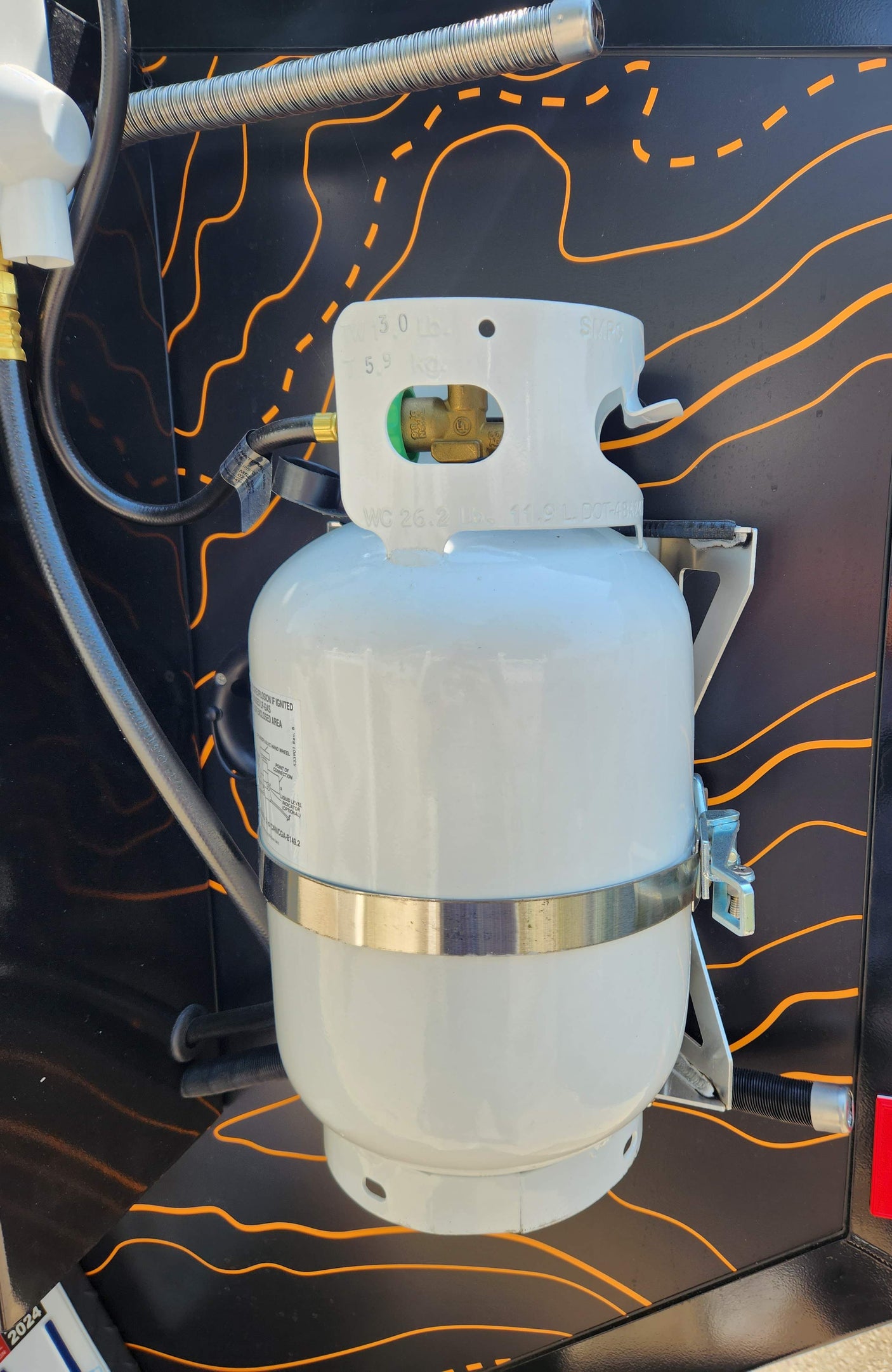 White propane tank with hoses, mounted on a black wall with orange lines in an RV rental setup.