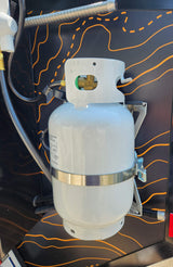 White propane tank with hoses, mounted on a black wall with orange lines in an RV rental setup.