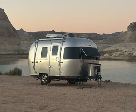 An Airstream RV rental camps by a serene lake at dusk, surrounded by rugged desert mountains, offering a perfect outdoor escape.
