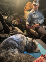 Man on phone, fluffy grey/white dog resting in cozy RV rental. Dashboard and accessories visible inside the camper.