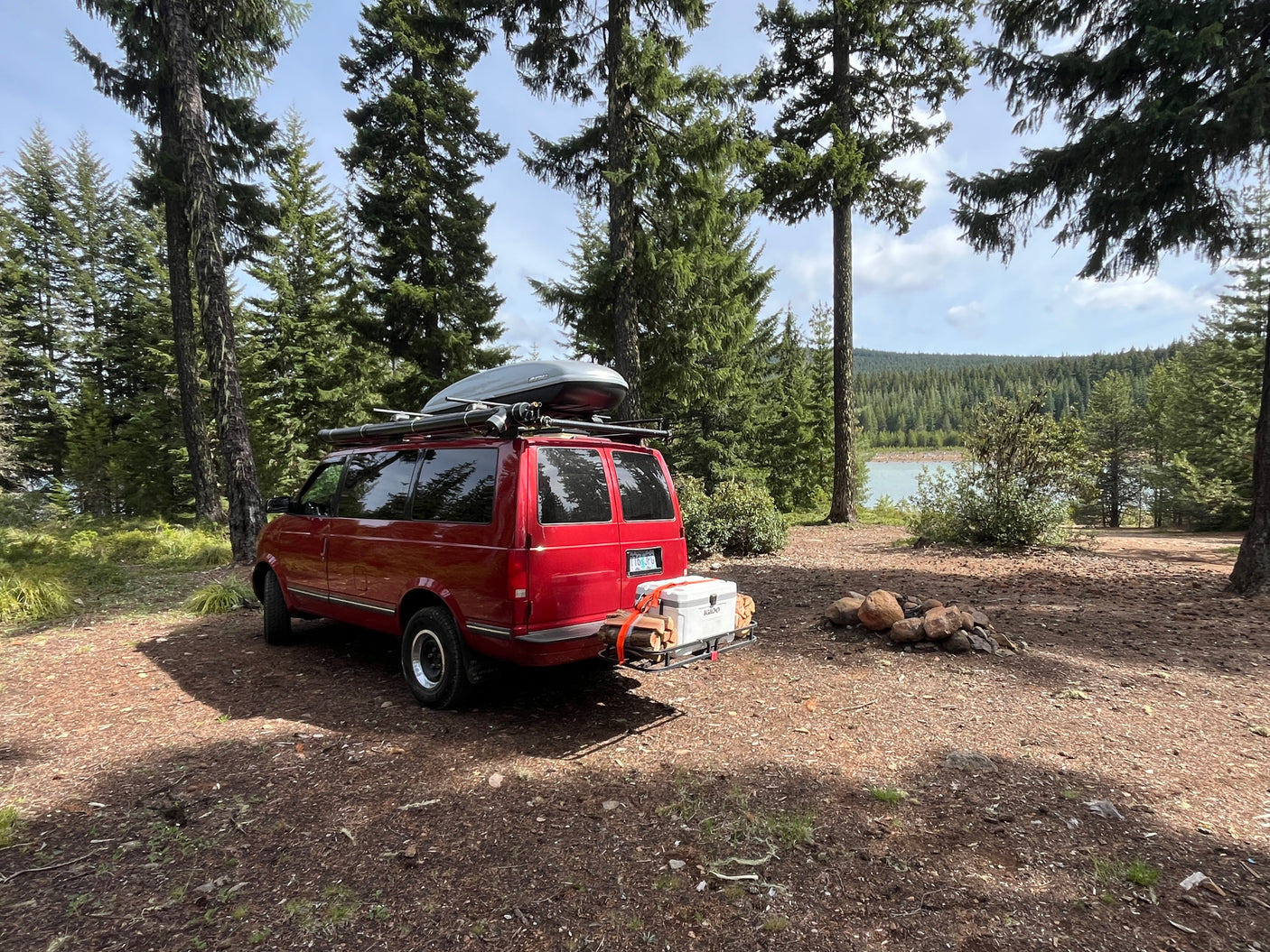 Camping with an rv rental! Red van parked by a lake in a forest, equipped with roof gear, cooler, and a fire pit.