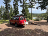 Camping with an rv rental! Red van parked by a lake in a forest, equipped with roof gear, cooler, and a fire pit.
