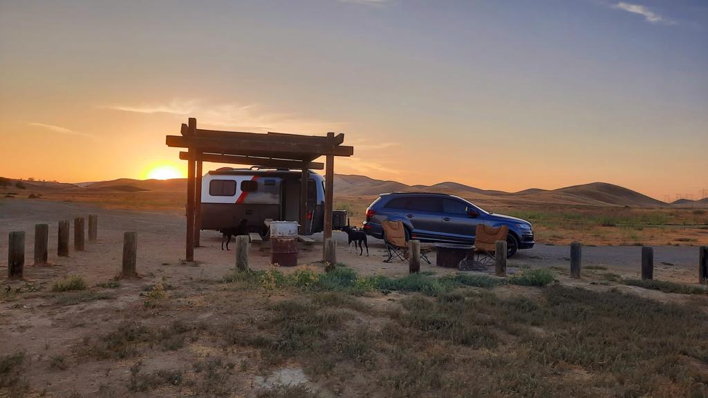 Sunset RV rental camping in arid hills: a small trailer under a pergola with an SUV, dogs, and fire pit.