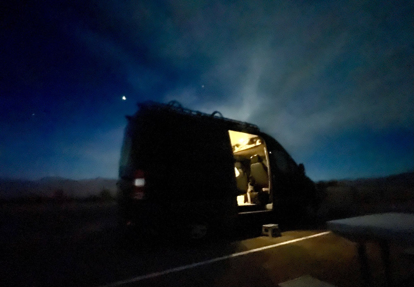 Nighttime rv rental: A camper van, door open revealing a warm interior, parked under a deep blue sky with a bright moon.