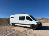 White camper van RV rental with bike on back, mat on ground, parked in sunny desert with hills.