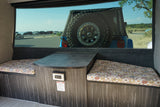 A cozy RV rental interior with patterned cushions, a table, and a window view of a blue Jeep parked near a beach.