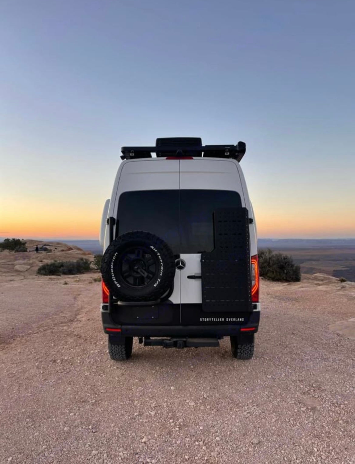 Rear view of a white Storyteller Overland adventure van (RV rental) with spare tire, parked on a dusty desert overlook at sunset.