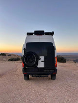 Rear view of a white Storyteller Overland adventure van (RV rental) with spare tire, parked on a dusty desert overlook at sunset.