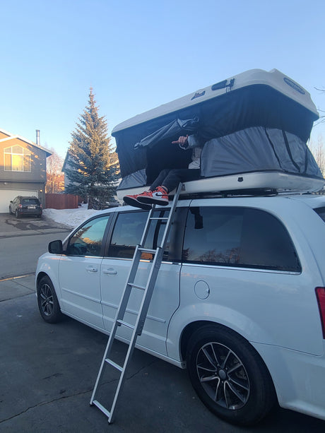 A person relaxes in a James Baroud rooftop tent on a white minivan, accessed by a ladder. A unique RV rental option for adventure.