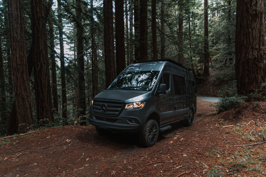 Dark grey Sprinter camper van, perfect for an rv rental, parked on a dirt path in a dense, towering forest.