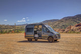 Custom grey Sprinter camper van, door open revealing interior, parked on dirt in mountains. Ideal for rv rental adventures.